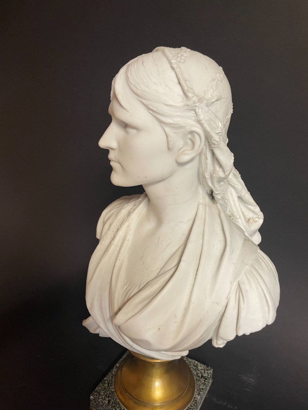 A white carved bust of a woman with draped classical clothing, mounted on a turned gold-tone socle and a square speckled granite base. View III
