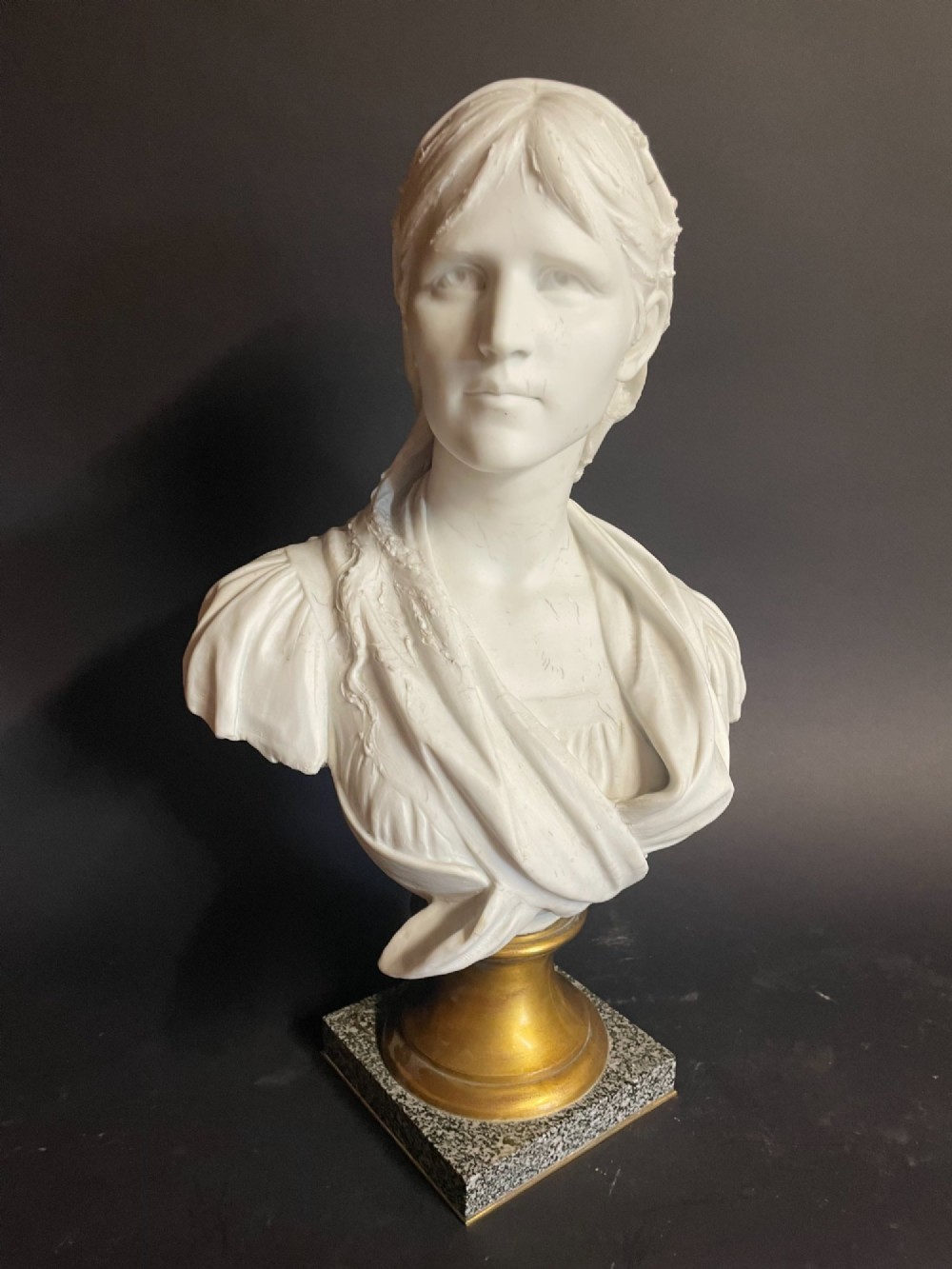 A white carved bust of a woman with draped classical clothing, mounted on a turned gold-tone socle and a square speckled granite base. View IV