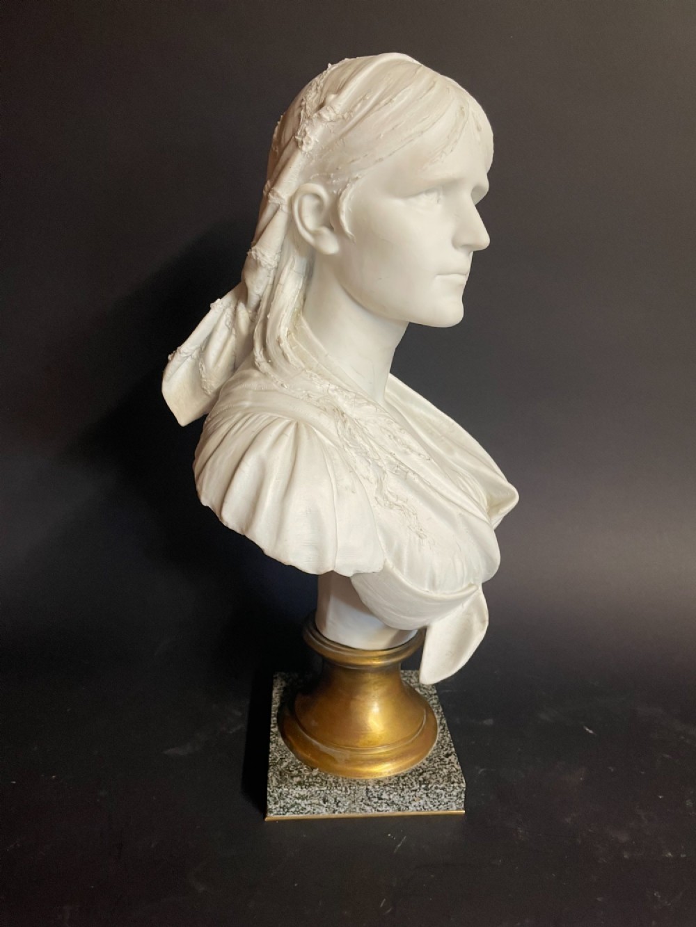 A white carved bust of a woman with draped classical clothing, mounted on a turned gold-tone socle and a square speckled granite base. View V