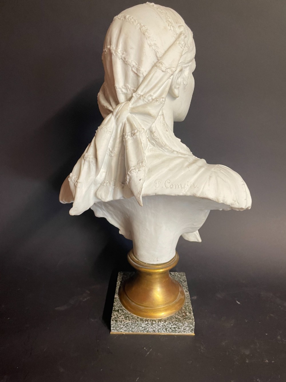 A white carved bust of a woman with draped classical clothing, mounted on a turned gold-tone socle and a square speckled granite base. View VI