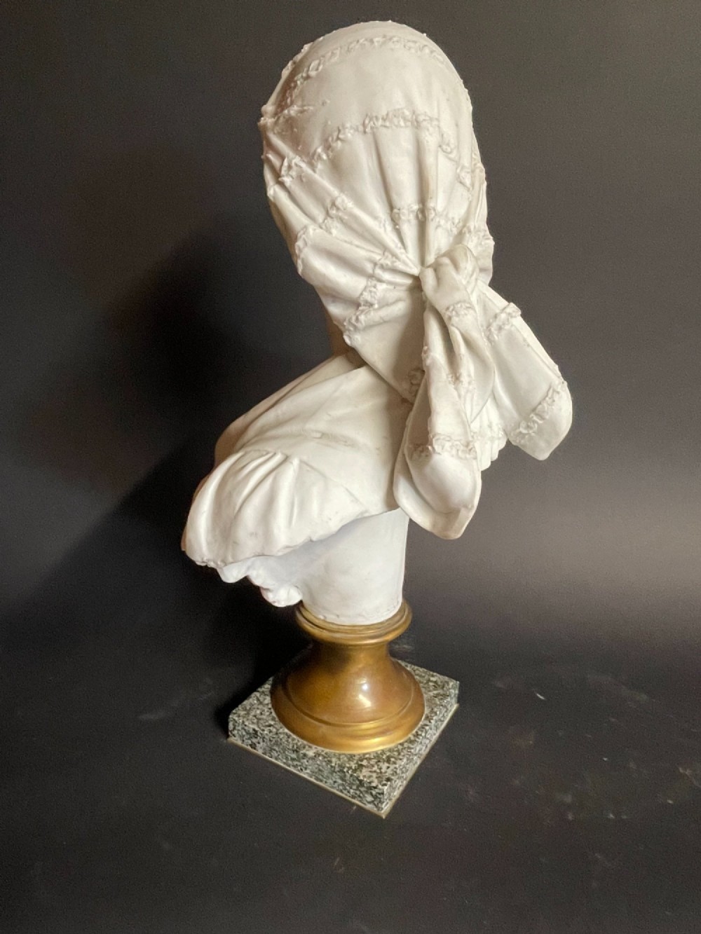 A white carved bust of a woman with draped classical clothing, mounted on a turned gold-tone socle and a square speckled granite base. View VIII