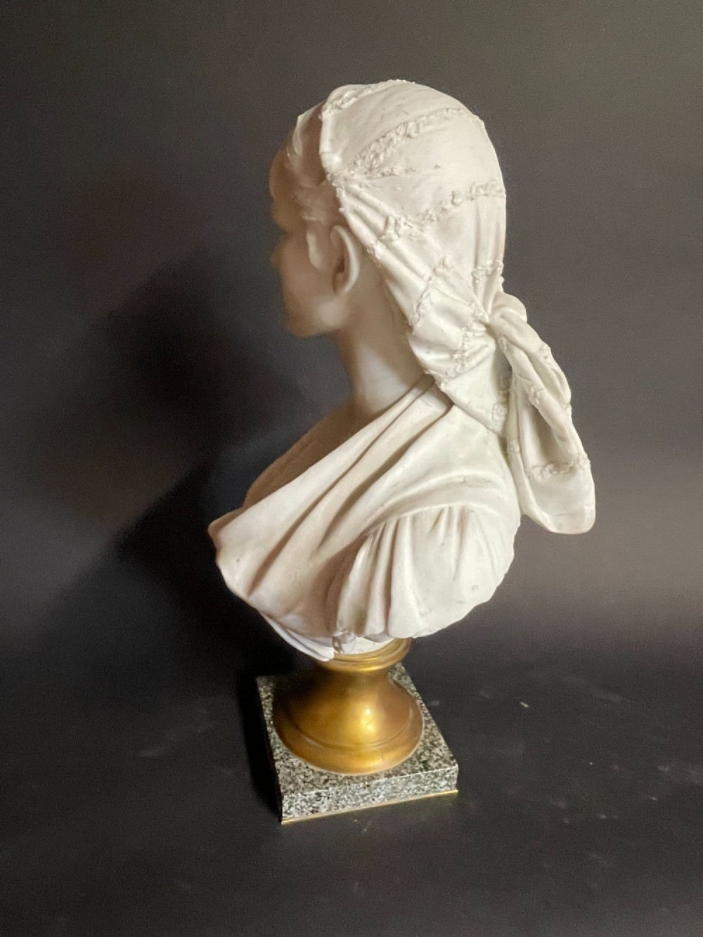 A white carved bust of a woman with draped classical clothing, mounted on a turned gold-tone socle and a square speckled granite base. View IX
