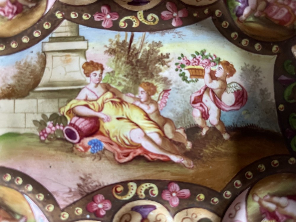 An oval enamelled dish with lobed edges, featuring hand-painted classical figures in the centre surrounded by cherub portraits and scrollwork along the border. View V