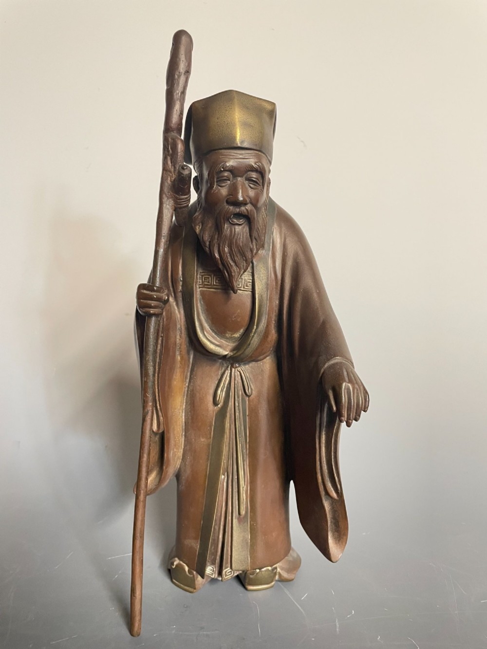 A patinated cast bronze Japanese figure depicting a bearded scholar wearing traditional robes and a tall headpiece while holding a walking staff.