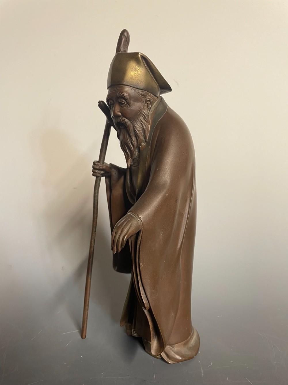 A patinated cast bronze Japanese figure depicting a bearded scholar wearing traditional robes and a tall headpiece while holding a walking staff. View III