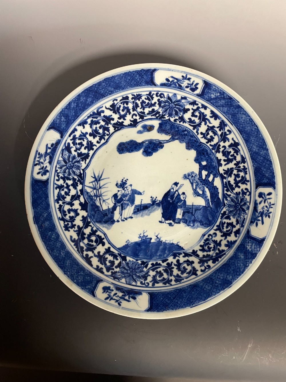A circular Chinese porcelain plate decorated with underglaze cobalt blue figures beneath a pine tree within floral and geometric patterned borders.