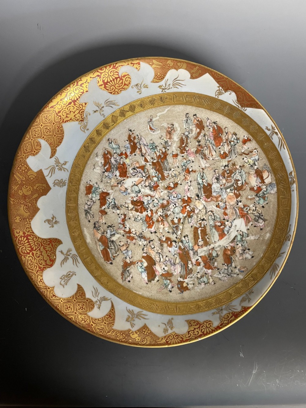 A circular Japanese Kutani porcelain dish featuring a central panel of hand-painted figures surrounded by an iron-red and gold patterned border.