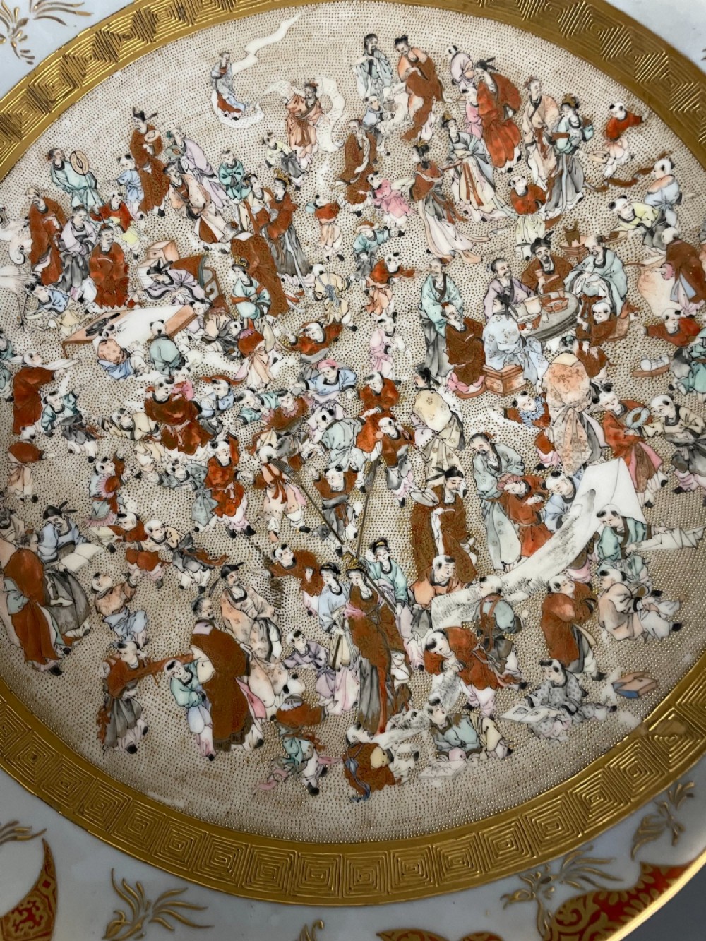 A circular Japanese Kutani porcelain dish featuring a central panel of hand-painted figures surrounded by an iron-red and gold patterned border. View II