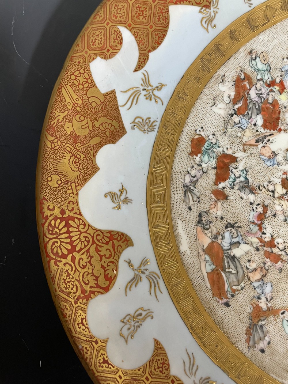 A circular Japanese Kutani porcelain dish featuring a central panel of hand-painted figures surrounded by an iron-red and gold patterned border. View III