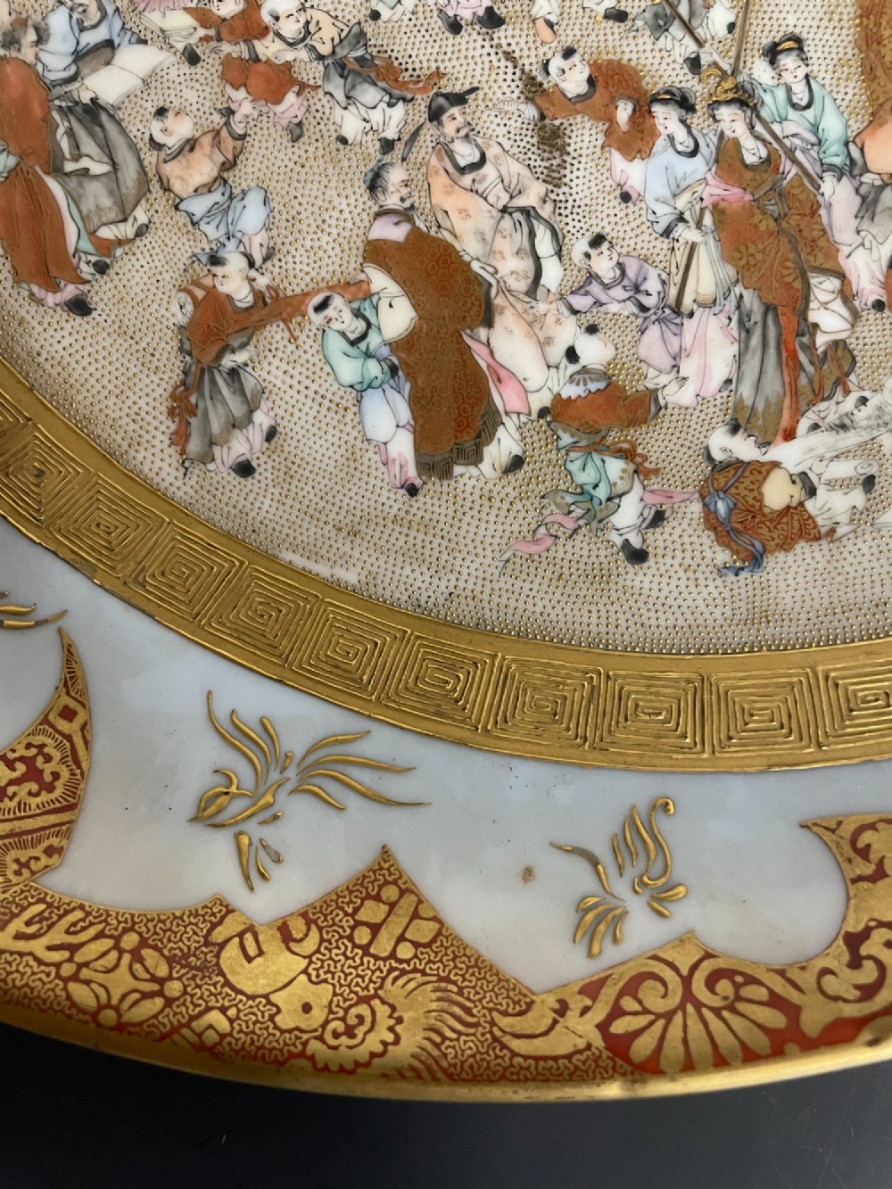 A circular Japanese Kutani porcelain dish featuring a central panel of hand-painted figures surrounded by an iron-red and gold patterned border. View IV