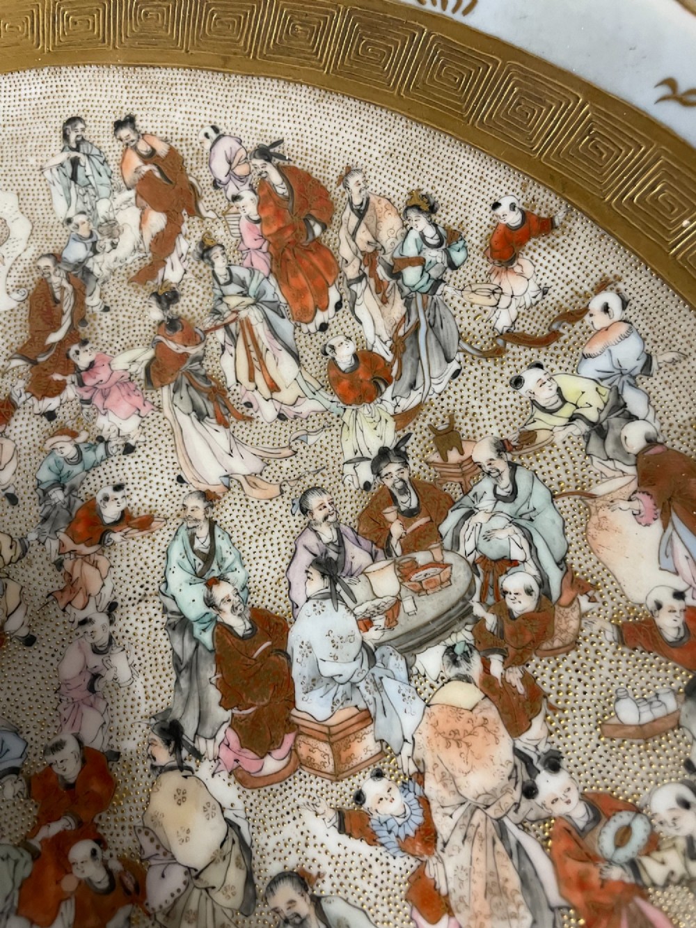 A circular Japanese Kutani porcelain dish featuring a central panel of hand-painted figures surrounded by an iron-red and gold patterned border. View V