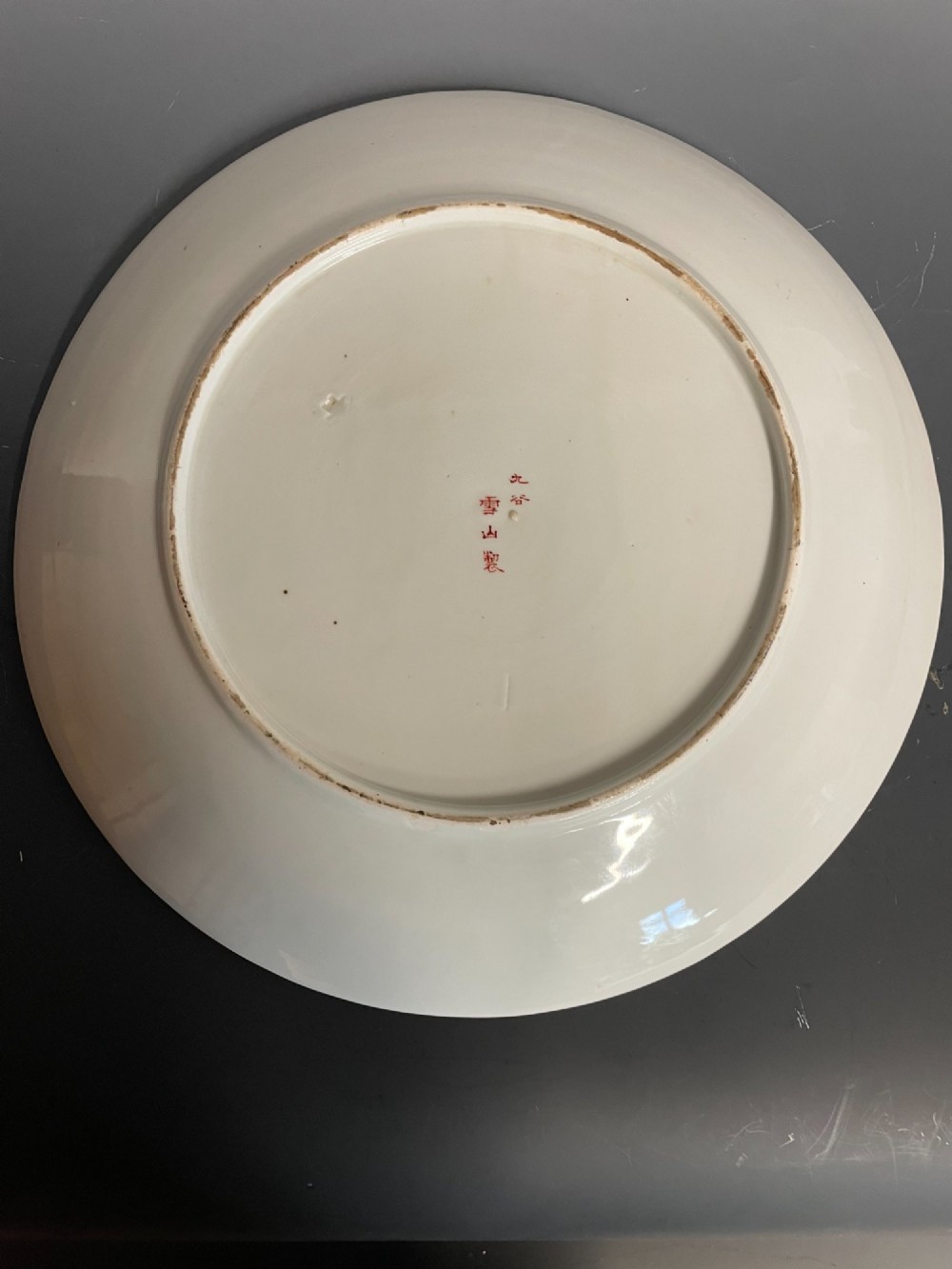 A circular Japanese Kutani porcelain dish featuring a central panel of hand-painted figures surrounded by an iron-red and gold patterned border. View VI