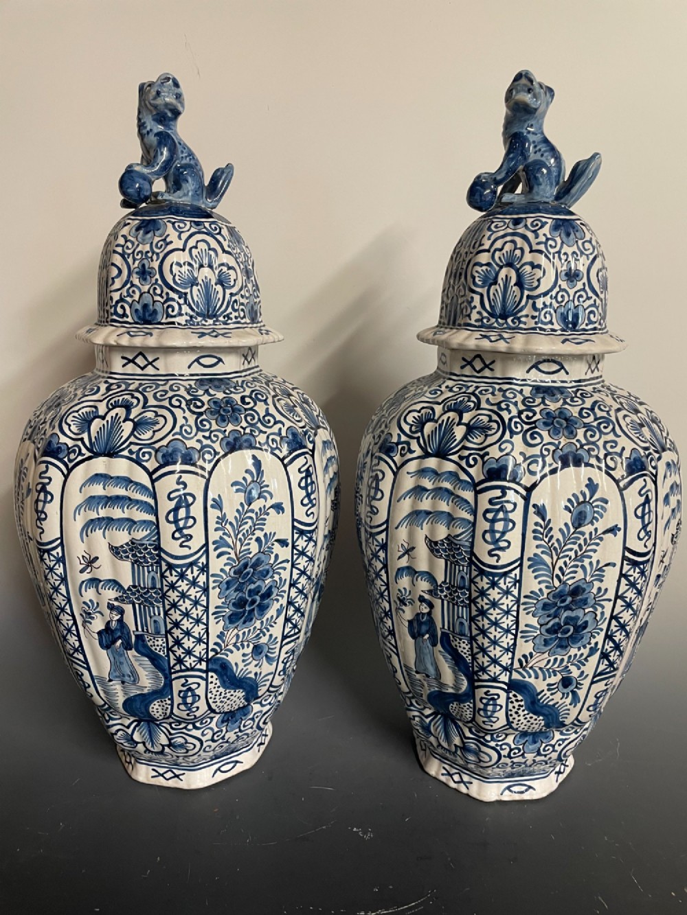 A pair of blue and white tin-glazed earthenware ribbed jars with lidded covers topped with foo dog finials, decorated with hand-painted chinoiserie scenes.