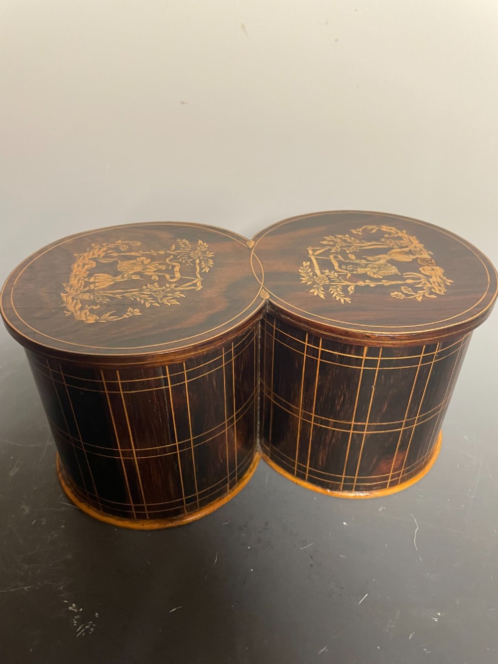 A dark brown wooden double cylindrical tea caddy featuring geometric line inlay and detailed figural marquetry scenes on the lids.