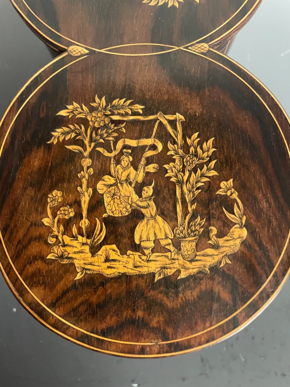 A dark brown wooden double cylindrical tea caddy featuring geometric line inlay and detailed figural marquetry scenes on the lids. View VI