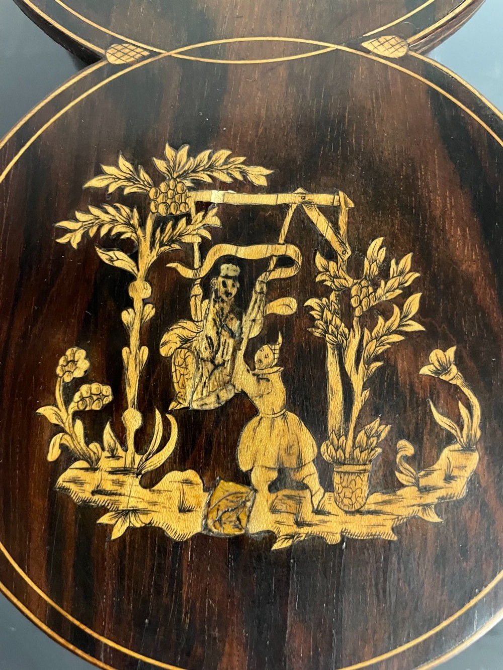 A dark brown wooden double cylindrical tea caddy featuring geometric line inlay and detailed figural marquetry scenes on the lids. View V