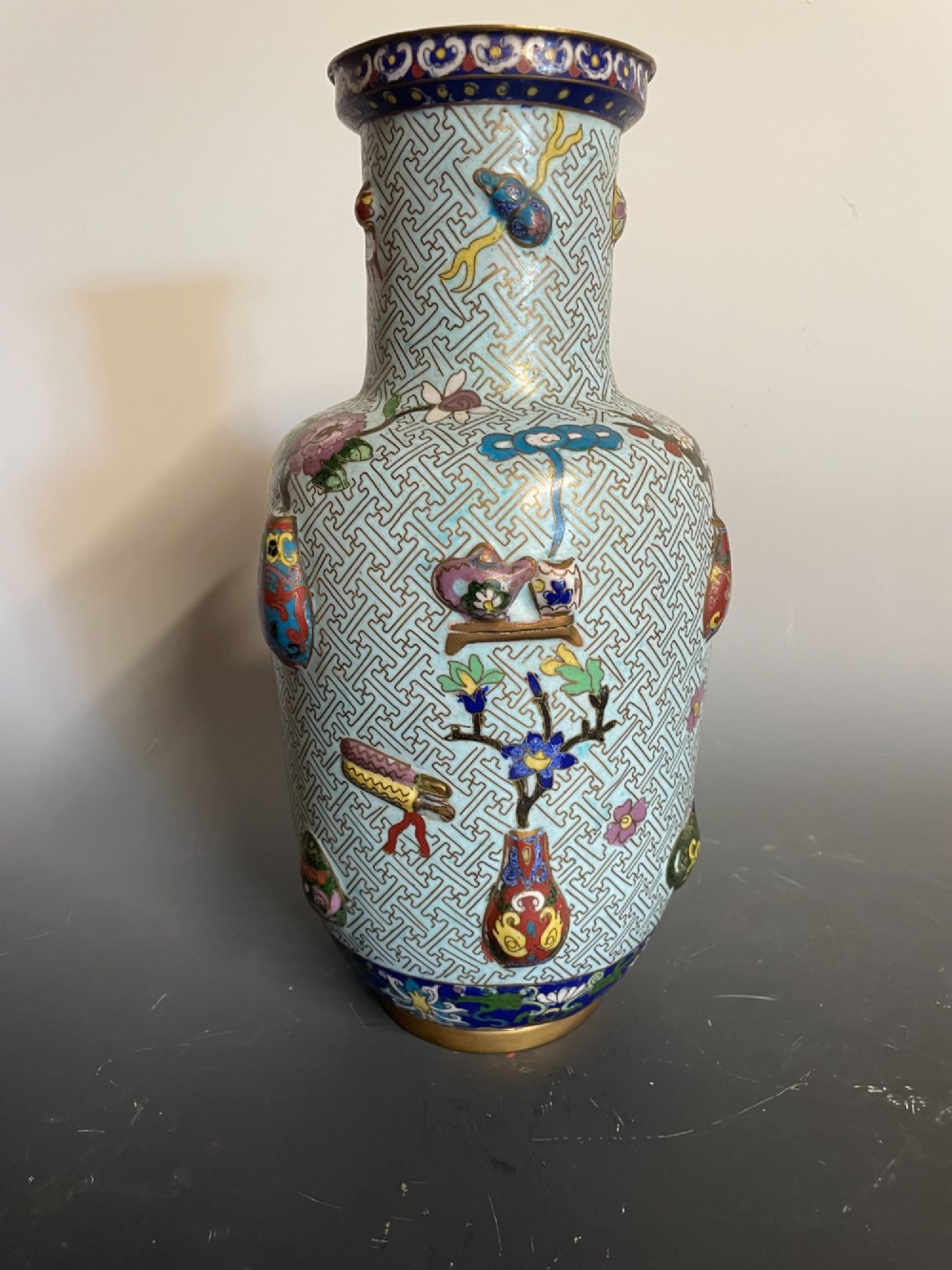 A baluster-shaped Chinese cloisonne enamel vase featuring a light blue key fret background embellished with colourful scholars objects and floral motifs.