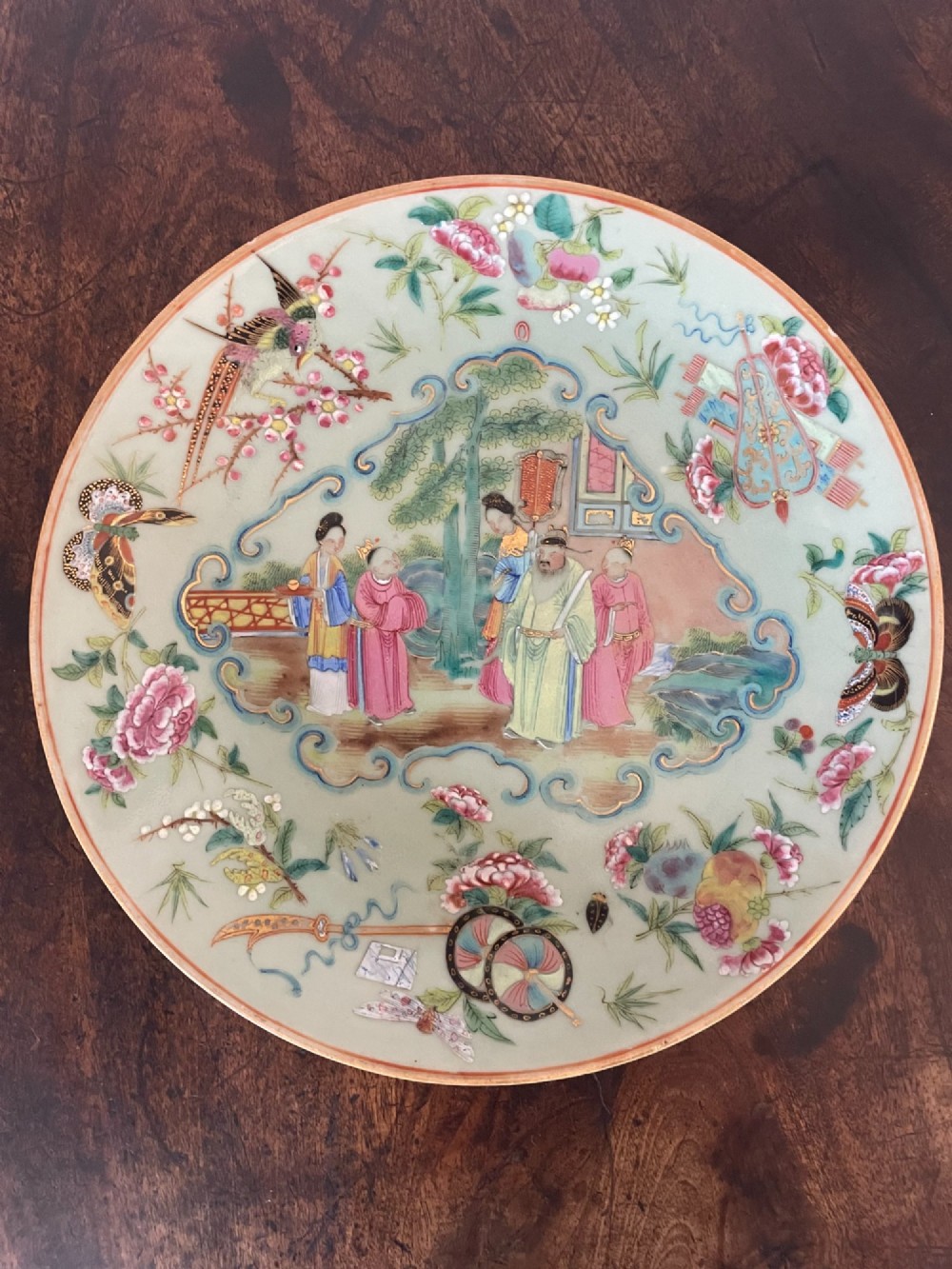 A circular Chinese export celadon porcelain plate hand-painted with a central figurative reserve surrounded by floral motifs, birds, and butterflies in multi-coloured enamels.