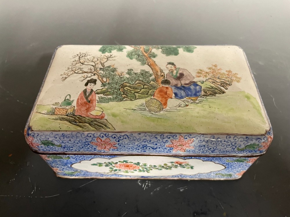 A rectangular Chinese Canton enamel box with rounded corners featuring hand-painted figures in a landscape setting on the lid and floral scrollwork on the side.