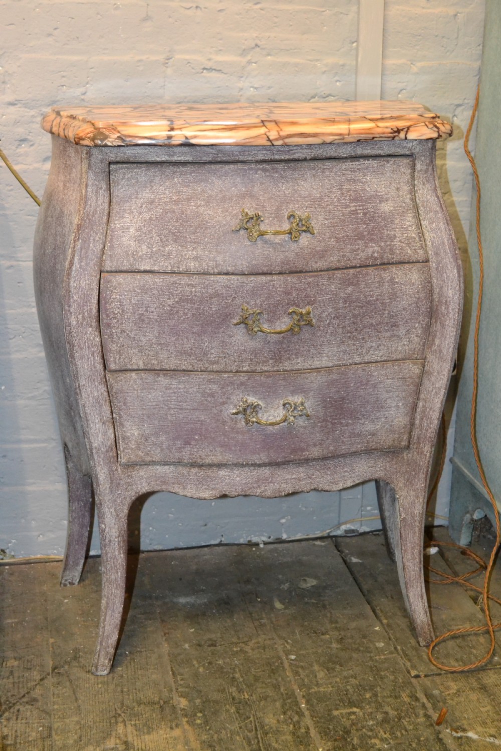A small French-style three-drawer commode with a distressed grey paint finish, curved cabriole legs, decorative brass handles, and a veined marble top.