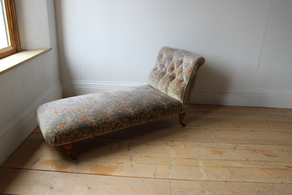 An upholstered antique day bed with a button-backed angled headrest, floral patterned grey fabric, turned wooden legs, and brass castors.
