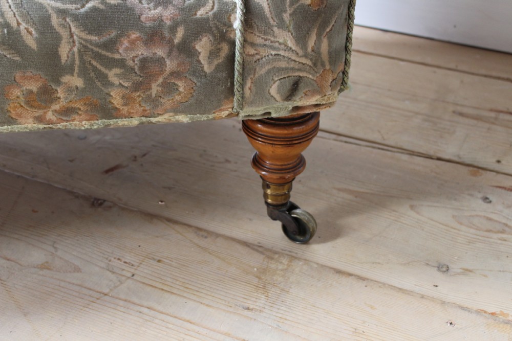 An upholstered antique day bed with a button-backed angled headrest, floral patterned grey fabric, turned wooden legs, and brass castors. View II