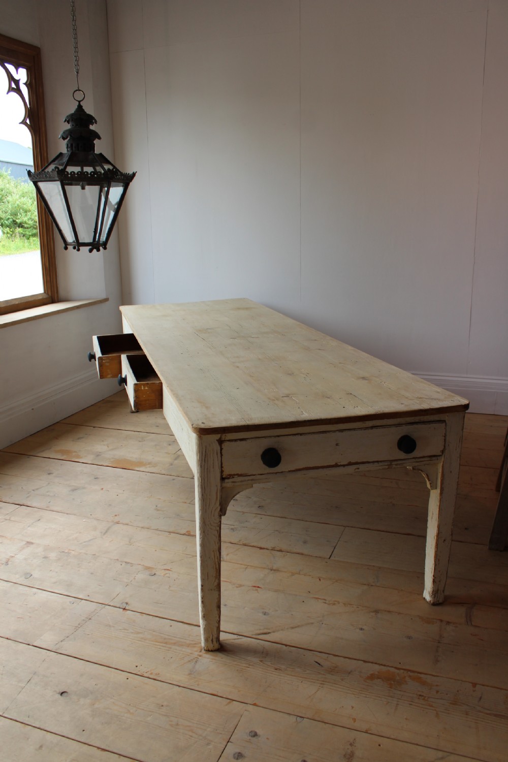 A long 19th-century country table with a natural scrubbed pine top, off-white painted base, and side drawers with dark wooden knobs.