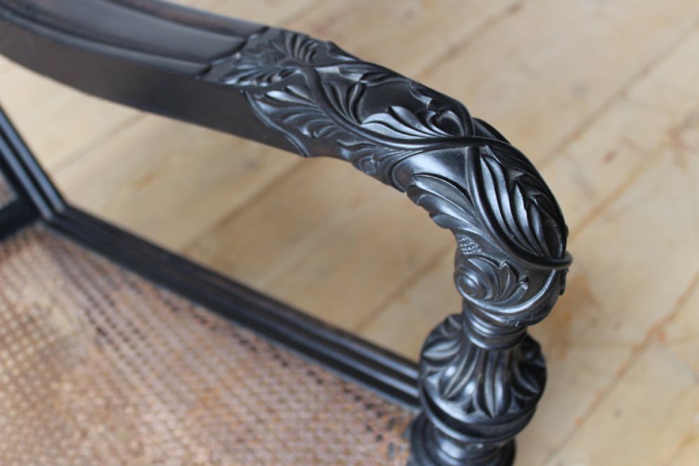 An antique dark carved wooden armchair featuring an intricately carved floral cresting rail, a woven cane backrest and seat, scrolled armrests, and turned legs on brass castors. View IV