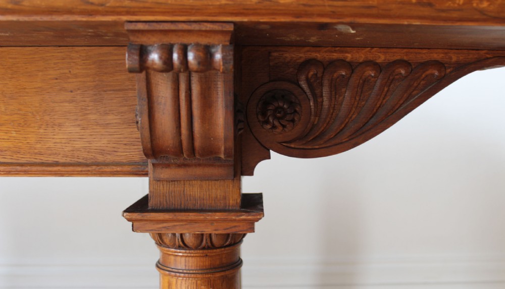 A 19th-century Arts and Crafts oak library table featuring four turned column legs, carved corner brackets, and a curved stretcher base. View IV