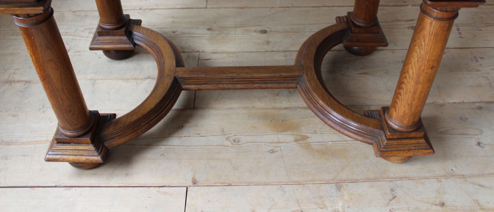 A 19th-century Arts and Crafts oak library table featuring four turned column legs, carved corner brackets, and a curved stretcher base. View VI