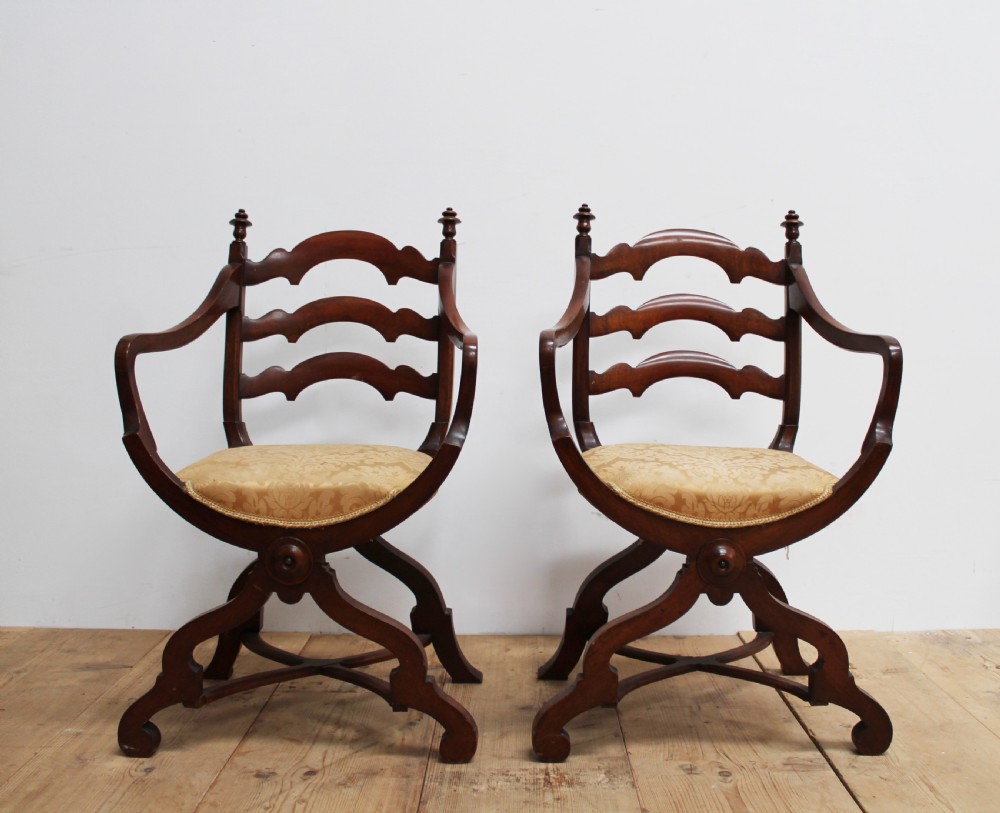 A pair of dark wood 19th-century X-frame armchairs featuring serpentine ladder-back splats, carved finials, and gold patterned fabric seats.