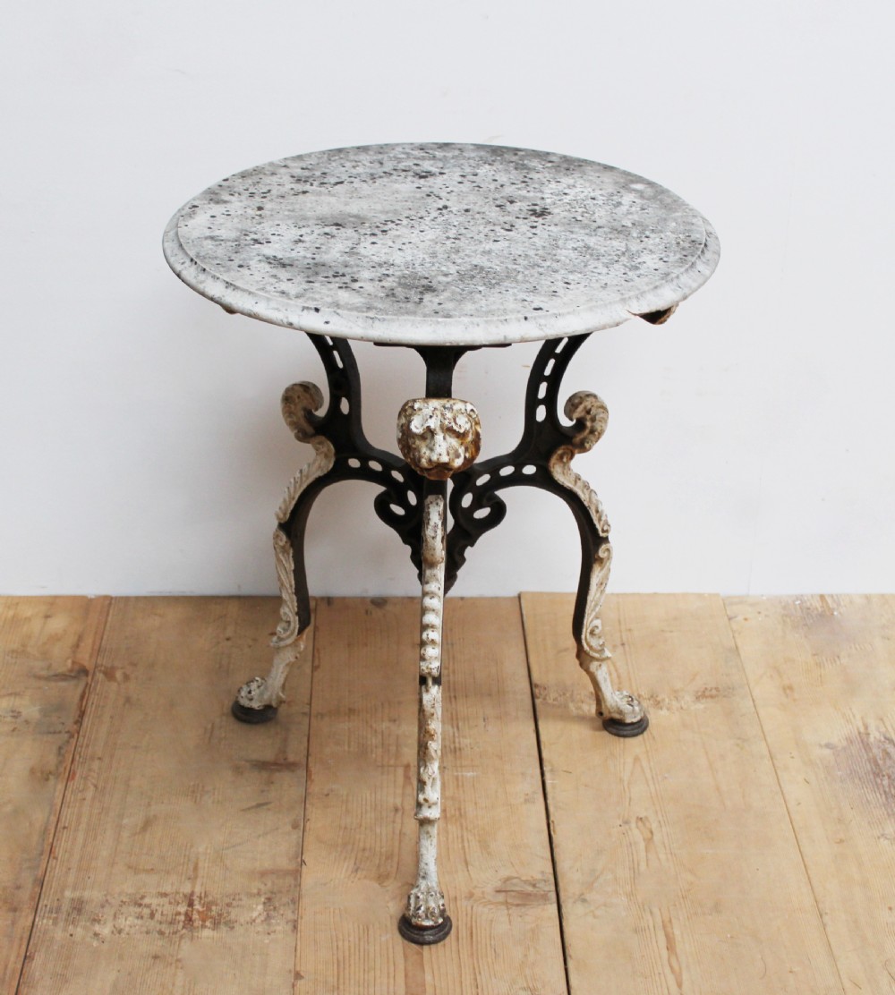 An antique nineteenth-century bistro table featuring a weathered circular marble top supported by a cast iron tripod base with lion mask details and claw feet.