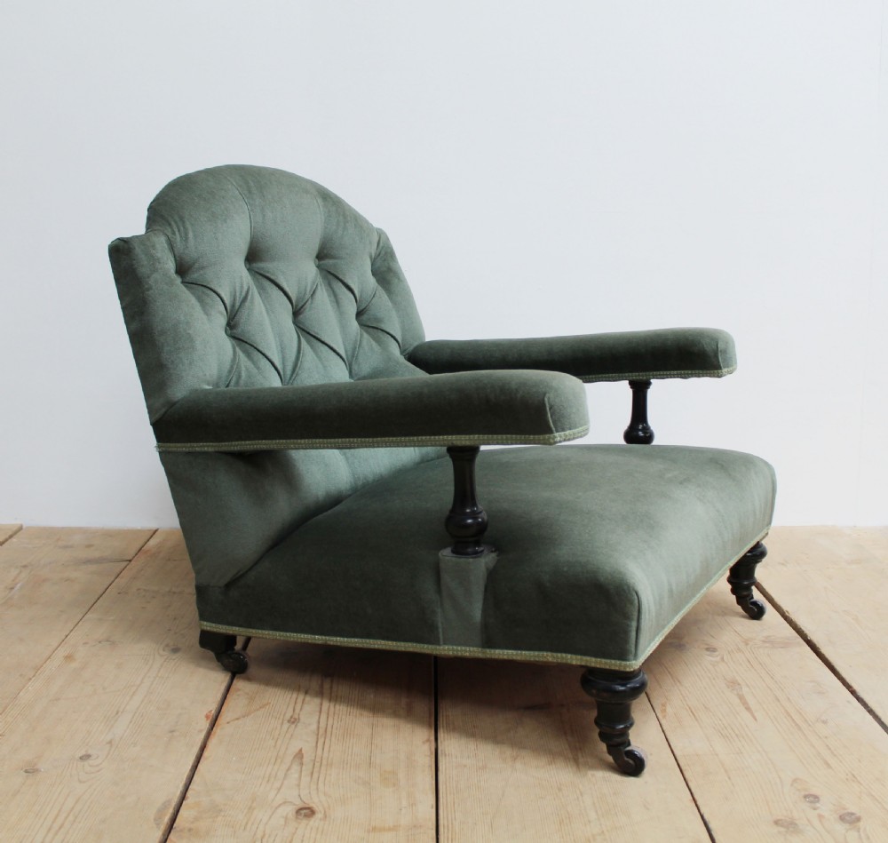 A low-slung Howard style armchair upholstered in sage green velvet featuring deep buttoning on the backrest and turned wooden legs on castors.