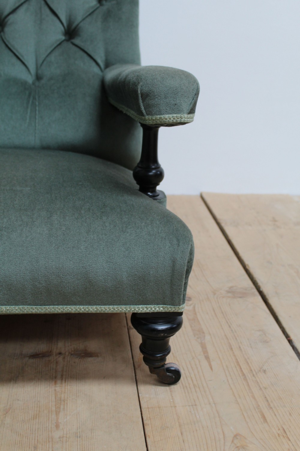 A low-slung Howard style armchair upholstered in sage green velvet featuring deep buttoning on the backrest and turned wooden legs on castors. View V