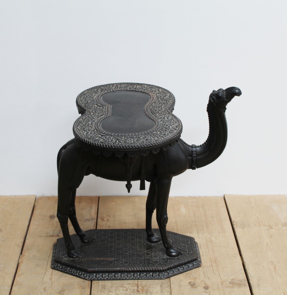 A dark carved wooden side table crafted in the form of a standing camel supporting a circular carved top on its saddle base. View II