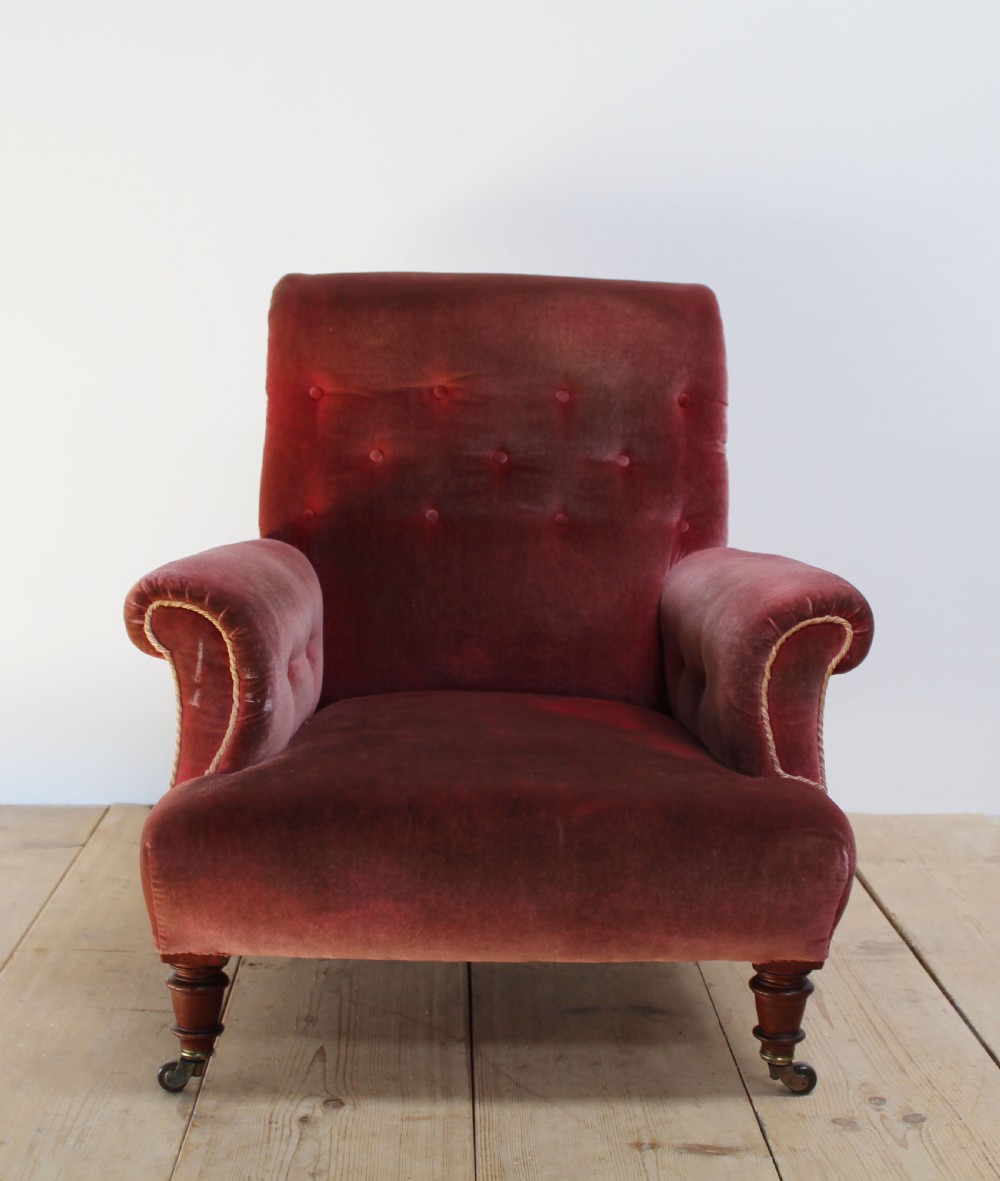 A Victorian armchair upholstered in faded red velvet featuring a button-backed backrest, rolled arms, and turned wooden front legs on brass castors.