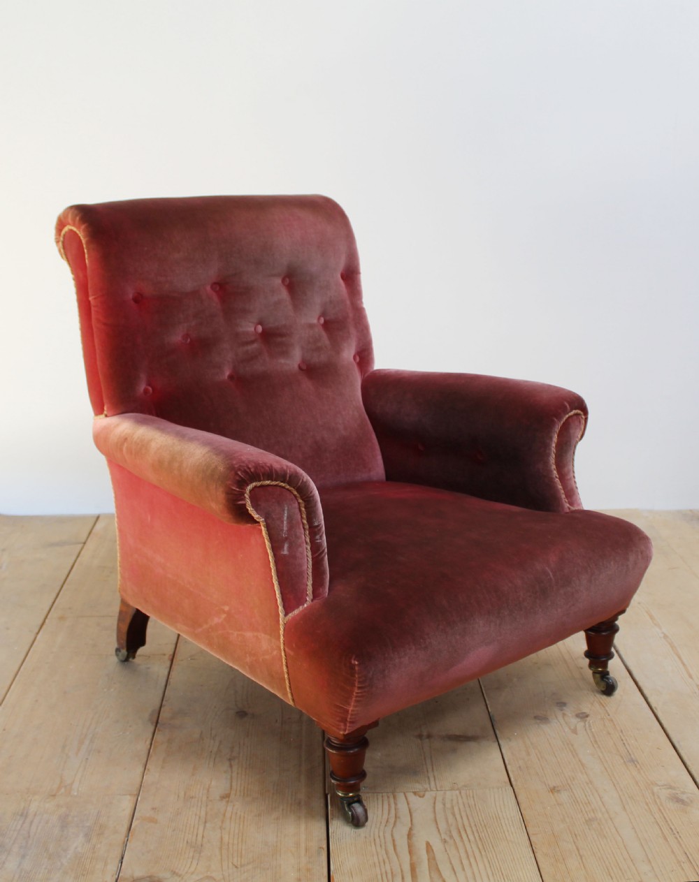 A Victorian armchair upholstered in faded red velvet featuring a button-backed backrest, rolled arms, and turned wooden front legs on brass castors. View IV