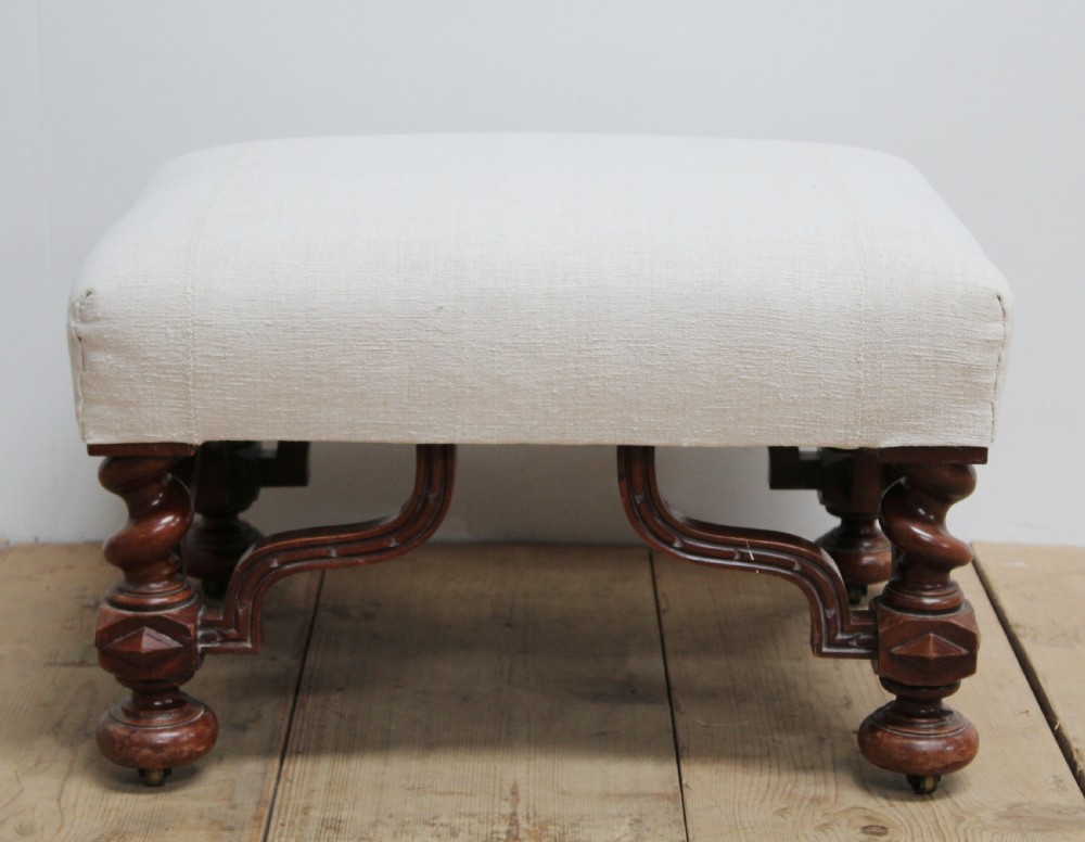 A low 19th-century footstool featuring cream linen upholstery, four carved barley twist legs, shaped stretchers, and brass castors.