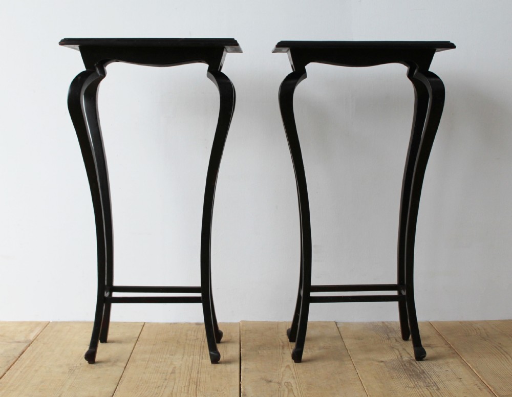 A pair of tall slender ebonised black wood console tables featuring outward-curving legs and square tops with lower stretchers.