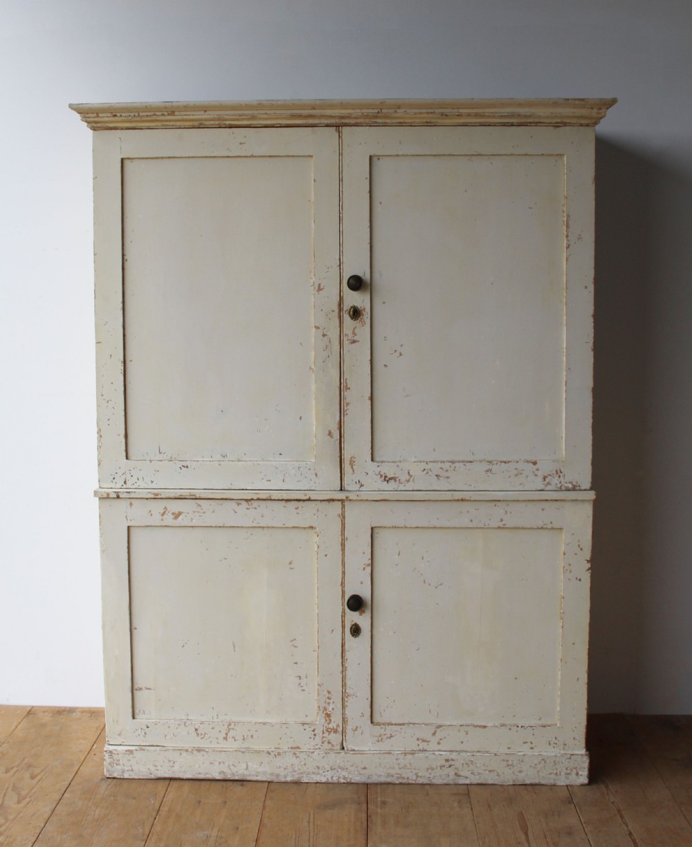 A tall 19th-century two-tier painted wooden linen cupboard with four panelled doors, a distressed off-white finish, turned wooden knobs, and brass keyhole escutcheons.