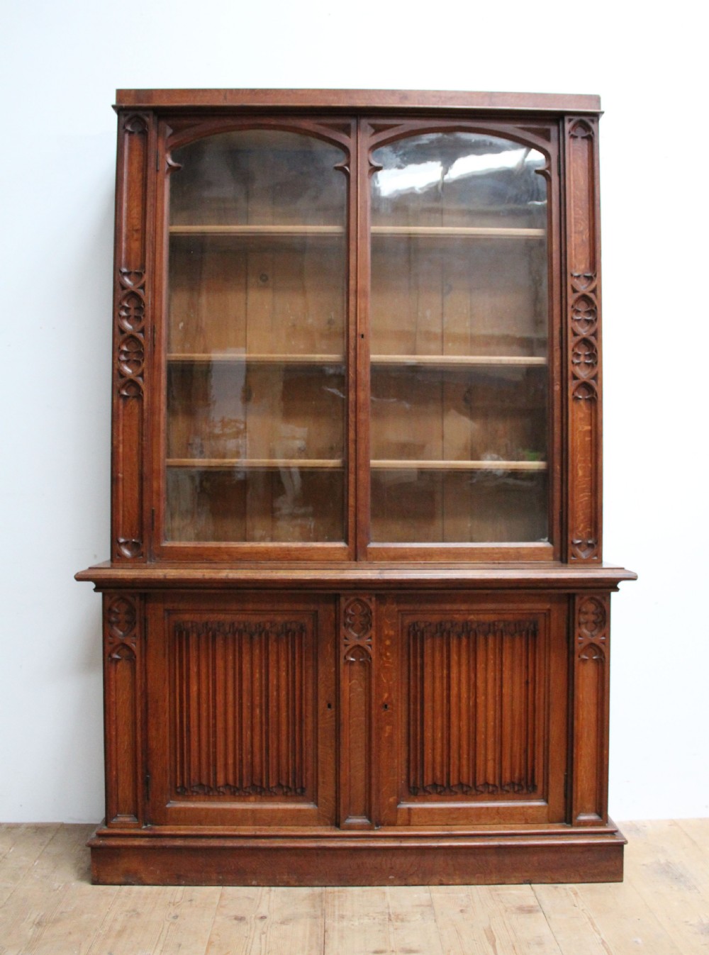 An Early Victorian Gothic Revival oak two-tier bookcase featuring double glazed upper doors with traceried arches and lower cupboard doors with carved linenfold panels.