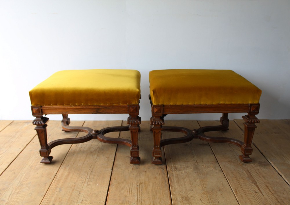 A pair of square upholstered stools featuring mustard yellow velvet seats and carved dark wood legs connected by curved cross-stretchers.