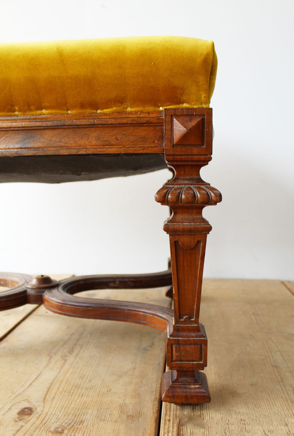 A pair of square upholstered stools featuring mustard yellow velvet seats and carved dark wood legs connected by curved cross-stretchers. View V