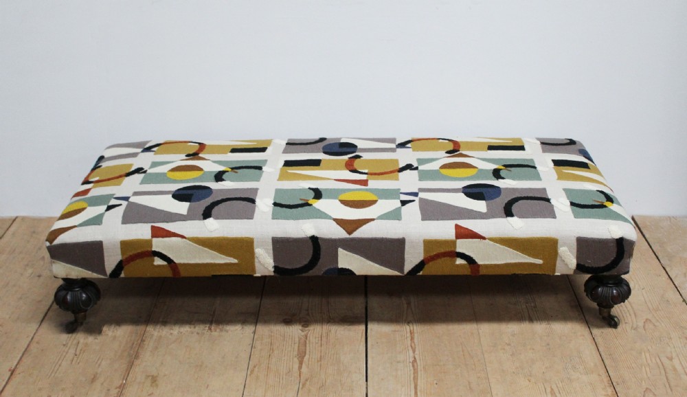 A large low rectangular ottoman upholstered in a geometric patterned textile, raised on four dark turned wooden legs with brass castors.