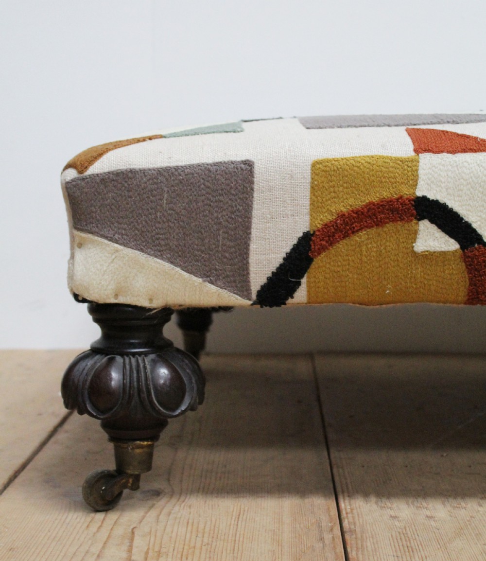 A large low rectangular ottoman upholstered in a geometric patterned textile, raised on four dark turned wooden legs with brass castors. View II
