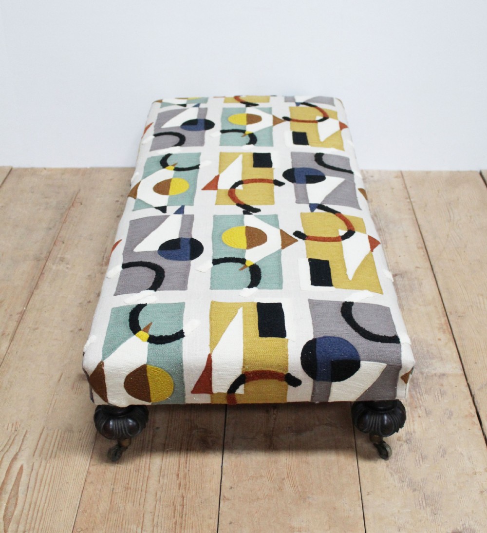 A large low rectangular ottoman upholstered in a geometric patterned textile, raised on four dark turned wooden legs with brass castors. View III
