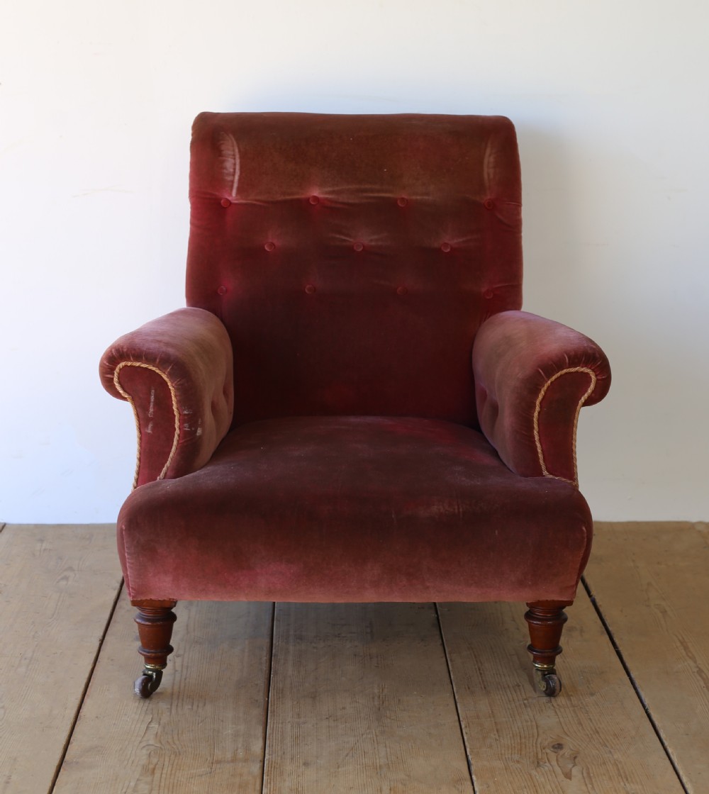 A 19th-century Victorian armchair upholstered in deep red velvet, featuring a buttoned backrest, scrolled arms with braid trim, and turned wooden front legs on brass castors.