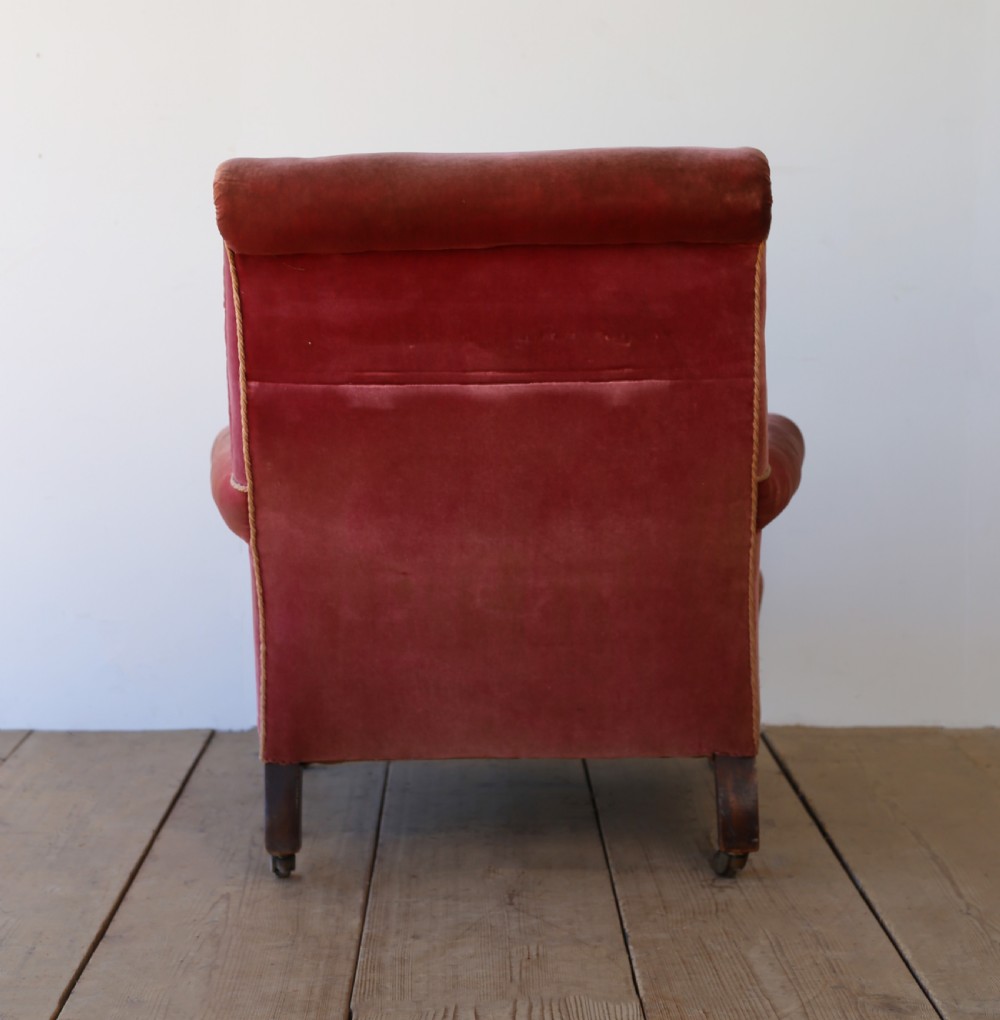A 19th-century Victorian armchair upholstered in deep red velvet, featuring a buttoned backrest, scrolled arms with braid trim, and turned wooden front legs on brass castors. View V