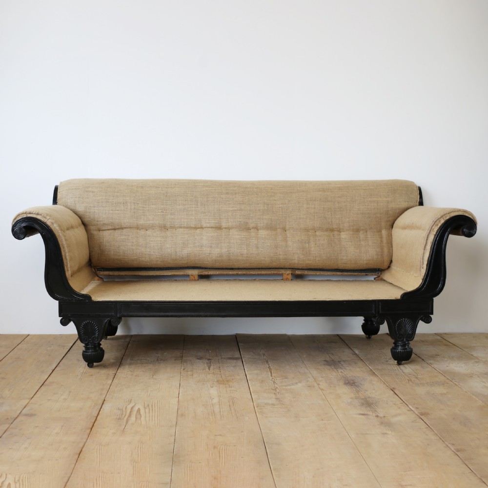A 19th-century Regency sofa featuring an ebonised dark wooden frame with scrolled arms, carved feet, and natural hessian lining.