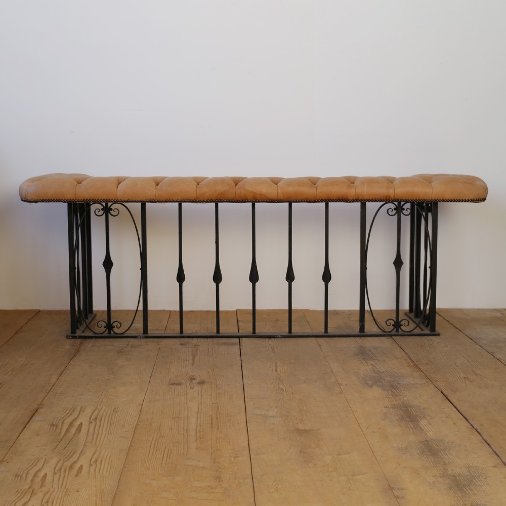 A 1920s English club fender featuring a buttoned tan leather bench top resting on a wrought iron baluster frame with scroll details.